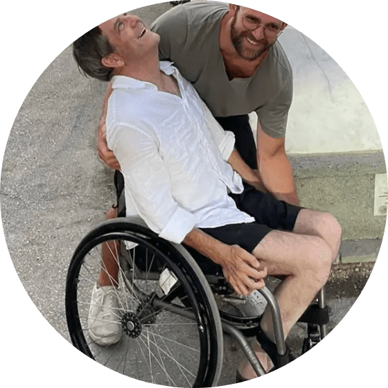Two men outdoors, one seated in a wheelchair, laughing together